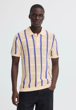 Jonge man draagt een beige gebreid poloshirt met verticale blauwe strepen, gecombineerd met zwarte broek, staand tegen een effen lichte achtergrond.