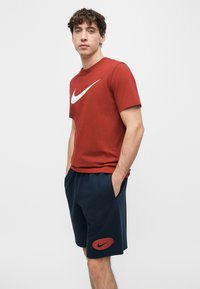 Νεαρός άνδρας που φοράει κόκκινο μπλουζάκι Nike και σκούρο μπλε σορτς Nike, όρθιος με τα χέρια στις τσέπες μπροστά σε ένα απλό λευκό φόντο.