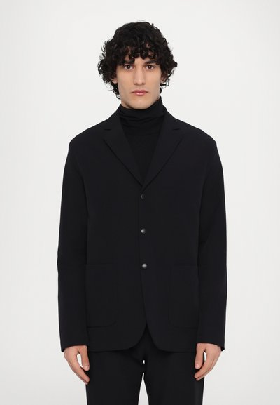 Schwarzer Blazer mit Reverskragen, zwei Fronttaschen und zwei Knöpfen. Getragen über einem schwarzen Rollkragenpullover, mit glatter Textur und klarer Struktur.