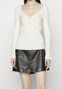 Maglietta a maniche lunghe avorio con dettaglio annodato sul petto, abbinata a una gonna a trapezio in pelle nera con finitura liscia.