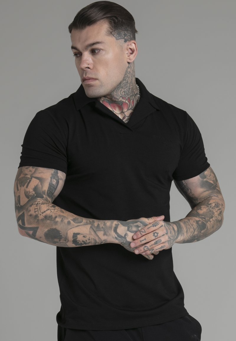 Juodas trumpomis rankovėmis polo marškinėliai su plokščiu apykakle. Modelis turi matomų tatuiruočių ant rankų ir kaklo, šukuosena suformuota, o jis stovi su sulenktais rankomis.