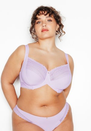 Krullendharige vrouw die een bijpassende lila bh en slipje met kanten rand draagt, staande tegen een witte achtergrond met haar handen achter haar rug.