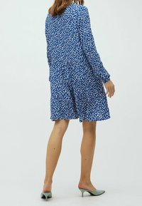 Ein blaues Kleid mit weißem Blattformmuster, langen Ärmeln, elastischem Bund und gestuften Rock. Model trägt grüne Mules und steht im Profil.