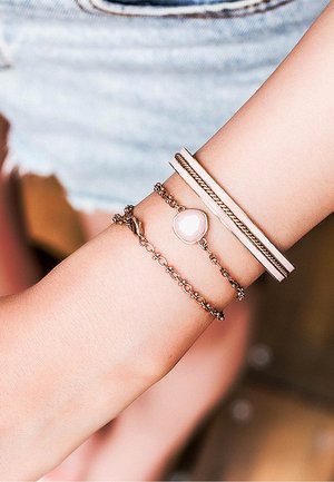 Quatre bracelets sur un poignet : une chaîne torsadée en or rose, une lanière métallique lisse, une chaîne délicate avec une pierre rose et une chaîne texturée.