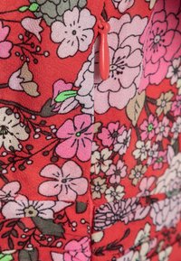 Tissu rouge avec un motif floral comprenant des fleurs roses et blanches. Comprend un zip rouge pour la fermeture. Texture douce et design vibrant.