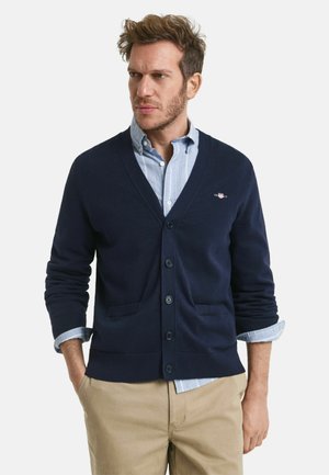 Cardigan en maille bleu marine avec un décolleté en V, cinq boutons, deux poches et des poignets côtelés, porté par-dessus une chemise rayée bleu clair.