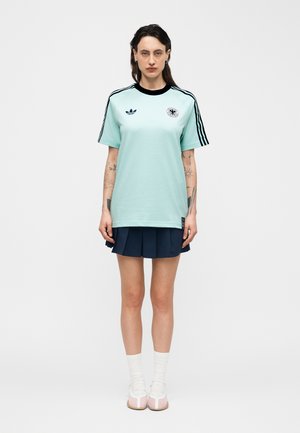 Moteris stovi, dėvi šviesiai mėlyną Adidas marškinėlį su juodomis juostelėmis ir logotipu, tamsiai mėlyną klostuotą sijoną, baltas kojines ir rožinius sportbačius.