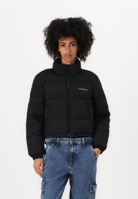 Zwarte pufferjack met een hoge kraag, ritssluiting en elastische manchetten. Voorzien van het 'GUESS JEANS' logo op de borst. Gestyled met blauwe denim broek.