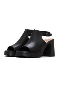Paire de sandales noires en cuir à bout ouvert, talon bloc, avec brides à la cheville et fermetures à boucle, sur fond blanc.