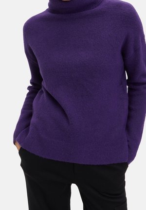 Person trägt einen lila Rollkragenpullover, hat die Hände in den Taschen einer schwarzen Hose und steht vor einem einfarbigen hellen Hintergrund.