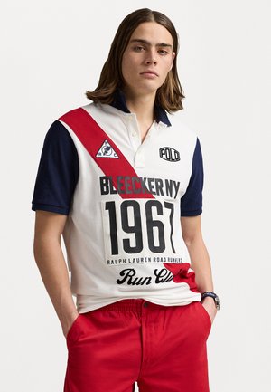 T-shirt polo blanc avec des manches bleu marine et une rayure diagonale rouge. Présente un grand texte graphique "1967" et des accents de logo. Associé à un short rouge.