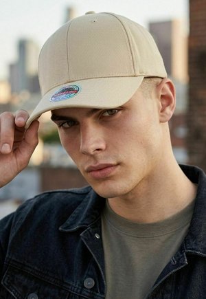 Young man wearing beige cap and dark denim jacket adjusts cap brim with cityscape blurred in background.