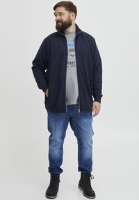 Veste zippée marine en tissu doux, avec des poches latérales, portée sur un T-shirt graphique gris, associée à un jean bleu et des bottes noires.
