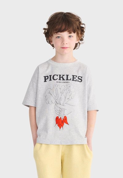 Kind mit lockigem Haar, das ein graues T-Shirt trägt, auf dem "PICKLES" steht, mit einer Hand, die blättriges Gemüse hält, und gelbe Hosen.