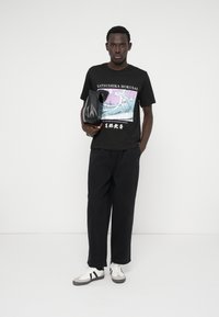 Svart t-shirt med en Hokusai-våg grafik, i kombination med svarta löst sittande byxor och vita sneakers med svarta ränder. Håller en svart väska.