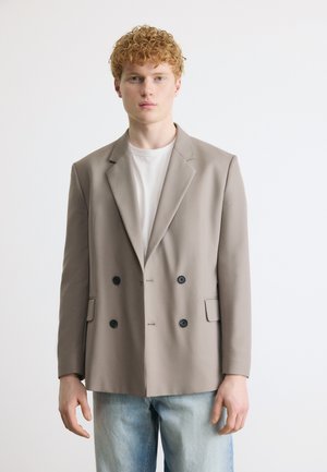 Mladý muž s kučeravými červenými vlasmi v béžovom dvojradovom saku, bielom tričku a svetlomodrých džínsach, stojaci pred bielym pozadím.