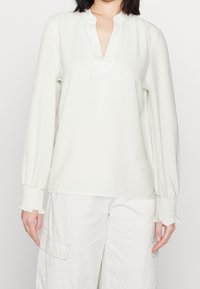 Weißes Blusenshirt mit V-Ausschnitt und langen, gerafften Ärmeln. Die Manschetten sind mit Smocking und subtilen Rüschendetails versehen. Weicher, leichter Stoff.
