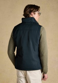 Mand iført mørkeblå polstret vest over olivengrøn sweater og lyse beige bukser, vendt væk mod neutral baggrund.