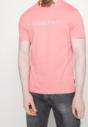 Person trägt ein pinkes Calvin Klein T-Shirt und dunkelgraue Jeans, steht mit entspannt an den Seiten hängenden Armen vor einem einfarbigen Hintergrund.