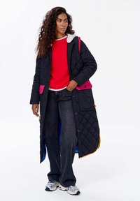 Žena s dlouhými kudrnatými vlasy, oblečená v černém prošívaném kabátu s růžovými kapsami, červeném svetru, černých džínech a stříbrných teniskách, stojící na bílém pozadí.