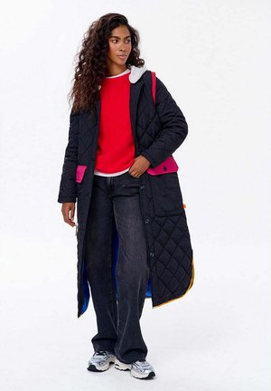 Žena s dlouhými kudrnatými vlasy, oblečená v černém prošívaném kabátu s růžovými kapsami, červeném svetru, černých džínech a stříbrných teniskách, stojící na bílém pozadí.