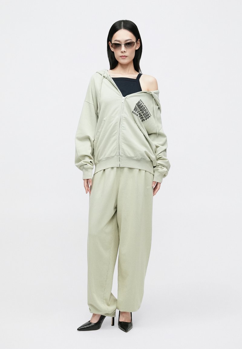 Lichtgroene zip-up hoodie met een grafische print, gecombineerd met bijpassende losse broek en een zwarte top. Model draagt strakke zwarte puntige hakken.