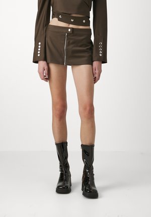 Brown faux leather mini skirt with a front zipper, paired with a cropped brown top featuring button accents and flared sleeves, worn with glossy black boots.