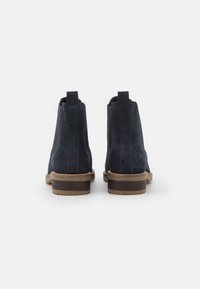 Un paio di stivali alla caviglia in suede blu scuro con tacco impilato marrone, mostrati da dietro su uno sfondo bianco.