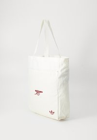 Weißer Canvas-Shopper mit zwei langen Griffen, vorderer Reißverschlusstasche, rotem Kanonengrafik und kleinem roten Adidas-Logo in der unteren Ecke.