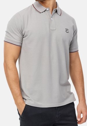Homme portant un polo gris clair à manches courtes avec col boutonné et petit logo brodé sur la poitrine, associé à un pantalon foncé.