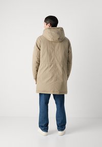 Beige parka berhood dengan finishing kasar, detail jahitan belakang, dan panjang hingga pertengahan paha. Dipadukan dengan celana jeans biru dan sepatu sneakers putih.
