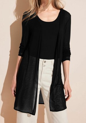 Vrouw draagt een lange zwarte open cardigan over een zwarte top en witte hoge taille broek tegen een neutrale achtergrond.