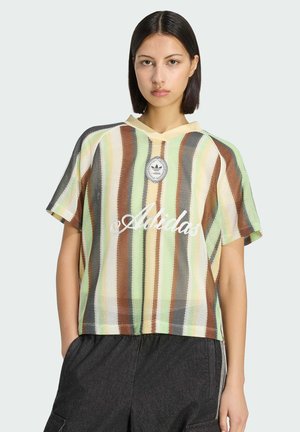 Donna con capelli lisci e scuri indossa una maglietta a maniche corte a righe con il logo Adidas e pantaloni larghi scuri, in posa su uno sfondo semplice.