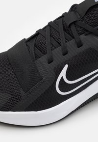 Zapato deportivo negro con parte superior de malla texturizada, cordones negros planos y un prominente logo blanco de Nike en el costado.
