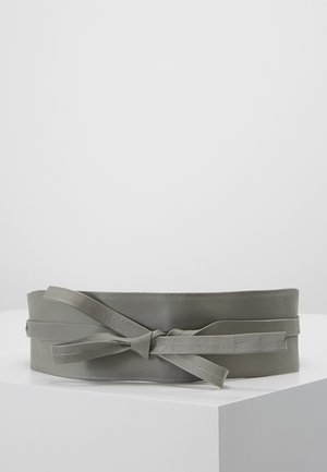 Leveä harmaa nahkavyö, solmittu rusetille, esillä valkoisella jalustalla yksiväristä vaaleanharmaata taustaa vasten.