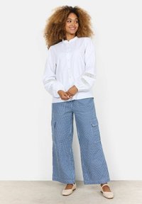 Camicia bianca con colletto arricciato e dettagli in pizzo sulle maniche, abbinata a pantaloni cargo a righe blu con gamba ampia. Scarpa di colore chiaro.