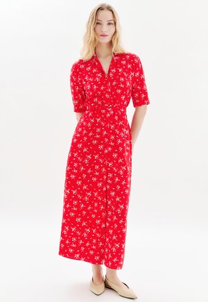 Robe fleurie rouge à manches courtes, avec un devant à boutons et une taille cintrée, fabriquée en tissu léger. Associée à des chaussures plates beiges.