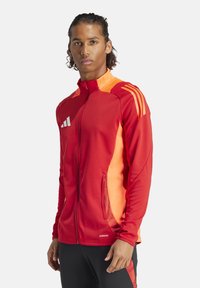 Rot-orange Sportjacke mit durchgehendem Reißverschluss, drei weißen Streifen auf der Schulter und seitlichen Mesh-Einsätzen, gefertigt aus leichtem Stoff.