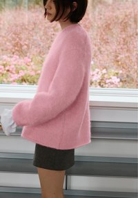 Femeie cu păr scurt și închis la culoare, purtând un pulover roz pufos și o fustă închisă la culoare, stând lateral lângă o fereastră cu flori roz în afară.