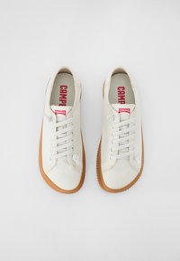 White canvas sneakers with beige rubber soles, featuring white laces, round toe, and a small logo on the side. Red branded insole.