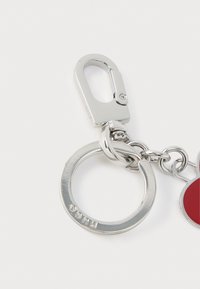 Porte-clés argenté avec un mousqueton, un anneau circulaire et un accent rouge. Présente une finition métallique polie et un logo en relief.