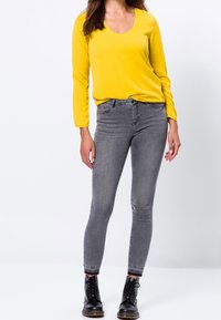 Gul langærmet top med rund hals, kombineret med grå skinny jeans. Sorte snørebåndsstøvler fuldender outfittet og fremhæver en tætsiddende silhuet.