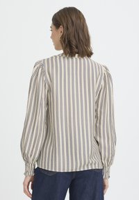 Blouse rayée beige clair avec des rayures bleu marine, manches bouffantes, col à volants et poignets froncés, associée à un jean denim bleu foncé.