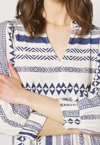 Vit klänning i tyg med blå geometriska mönster, med V-ringning, långa ärmar och draperade detaljer vid ärmsluten.