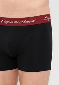 Boxers noirs avec une taille élastique bordeaux portant les inscriptions "Originals Studio" en écriture blanche. Tissu lisse avec des coutures contrastantes.