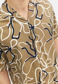 Chemise à manches courtes beige avec des motifs floraux abstraits bleu marine et blancs. Dotée d'un col classique et de boutons à l'avant.