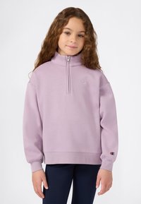 Jeune fille aux cheveux bruns bouclés, portant un sweat à col zippé pastel violet et un pantalon sombre, se tenant devant un fond uni.