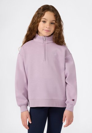 Jeune fille aux cheveux bruns bouclés, portant un sweat à col zippé pastel violet et un pantalon sombre, se tenant devant un fond uni.
