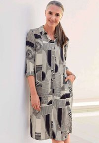 Knopfkleid aus leichtem Stoff mit einem geometrischen schwarz-cremefarbenen Muster, mit Kragen und hochgekrempelten Ärmeln. Mit seitlichen Taschen.