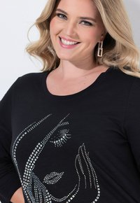 Lächelnde Frau mit welligem blonden Haar, die rechteckige Ohrringe und ein schwarzes Shirt mit einem funkensprühenden abstrakten Gesichtsmuster trägt.